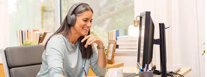 Smiling woman with headphones sitting at a desk, looking at a computer screen during a video call or online meeting.