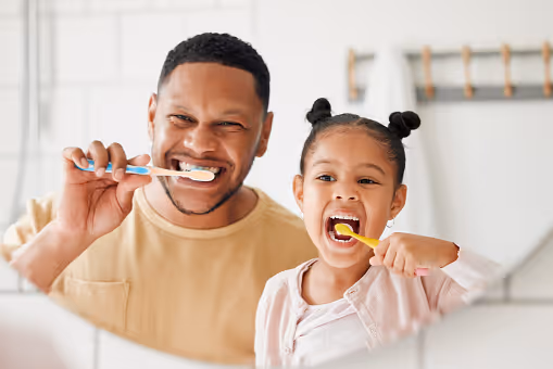 Father and daughter smiling together while brushing their teeth in a bathroom mirror.