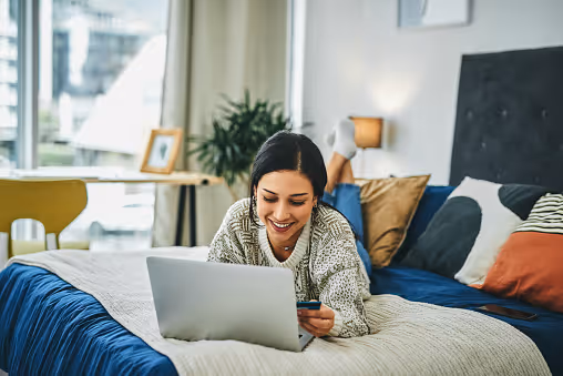Woman lying on bed using laptop and holding a credit card, smiling in a cozy bedroom.