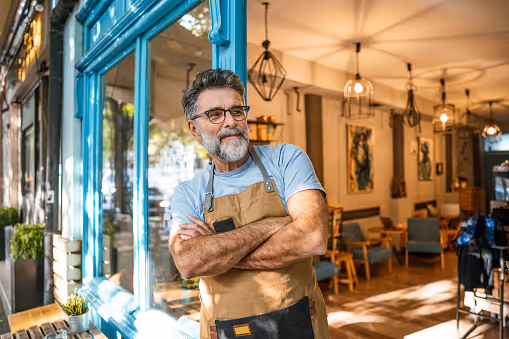 Smiling man in glasses and apron standing with arms crossed in front of a bright cafe window.