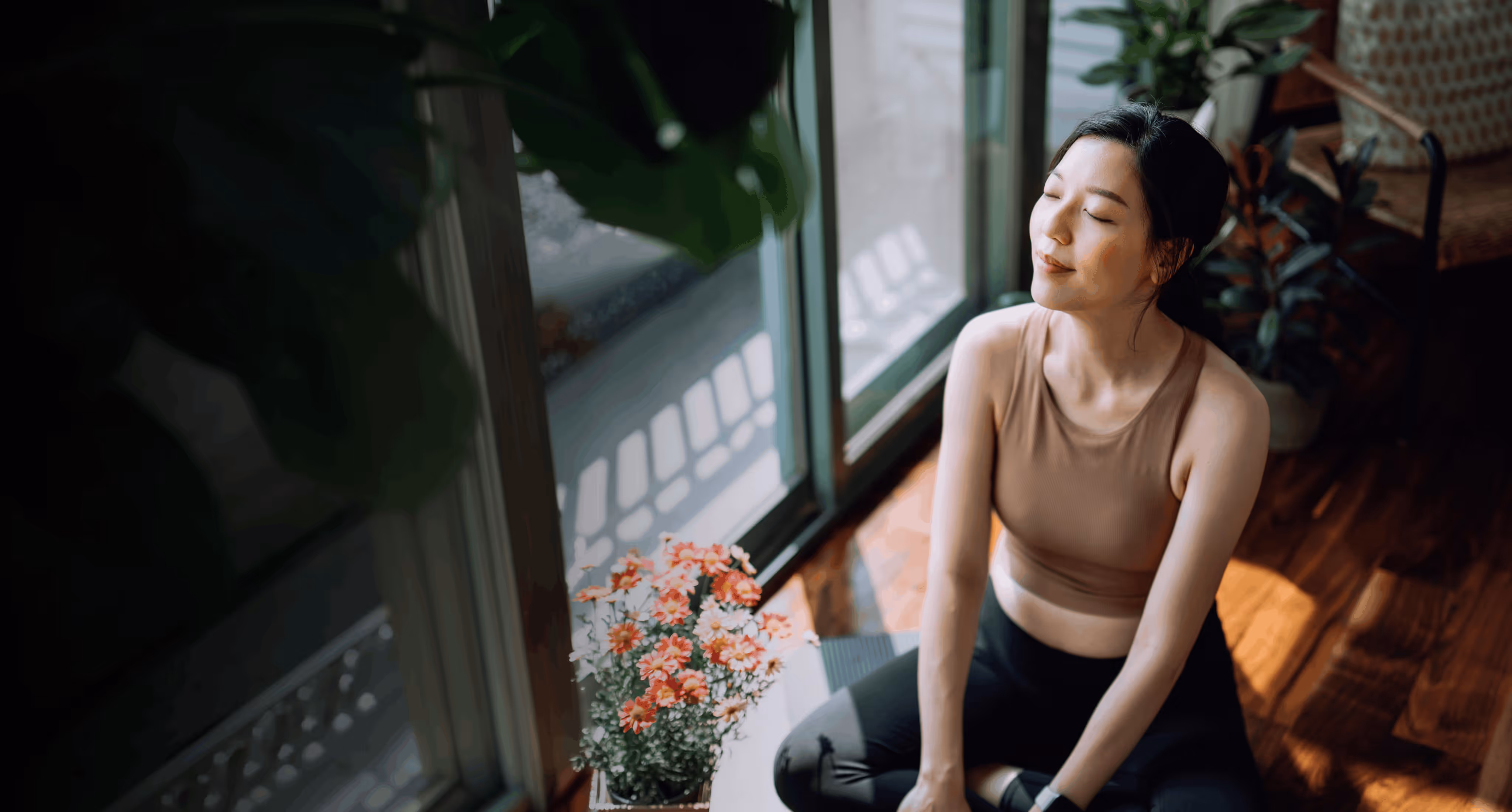 Woman in workout clothes sitting by window with eyes closed, enjoying sunlight near pink flowers.