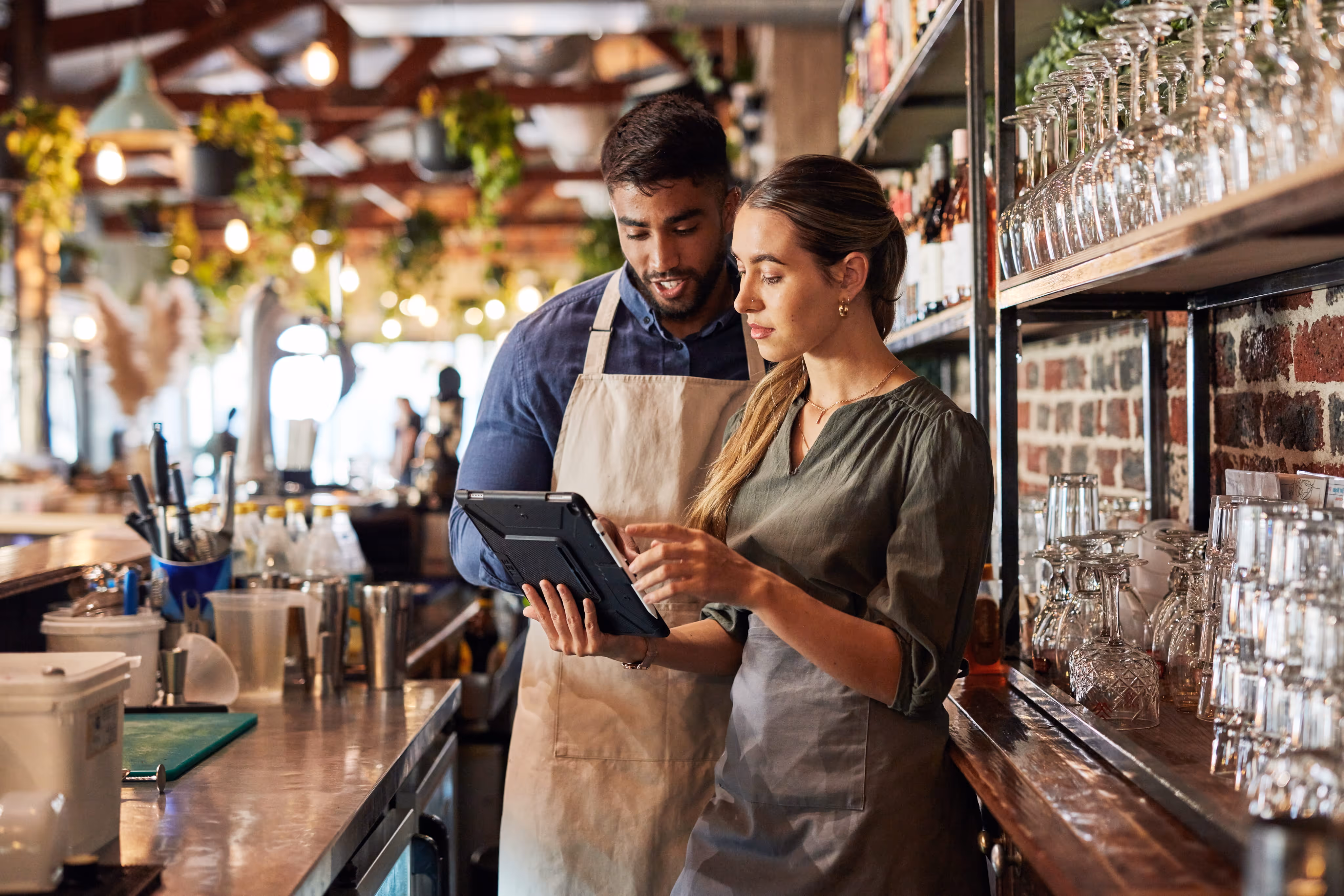 Two bar staff wearing aprons looking at a tablet together behind a bar counter with glasses.