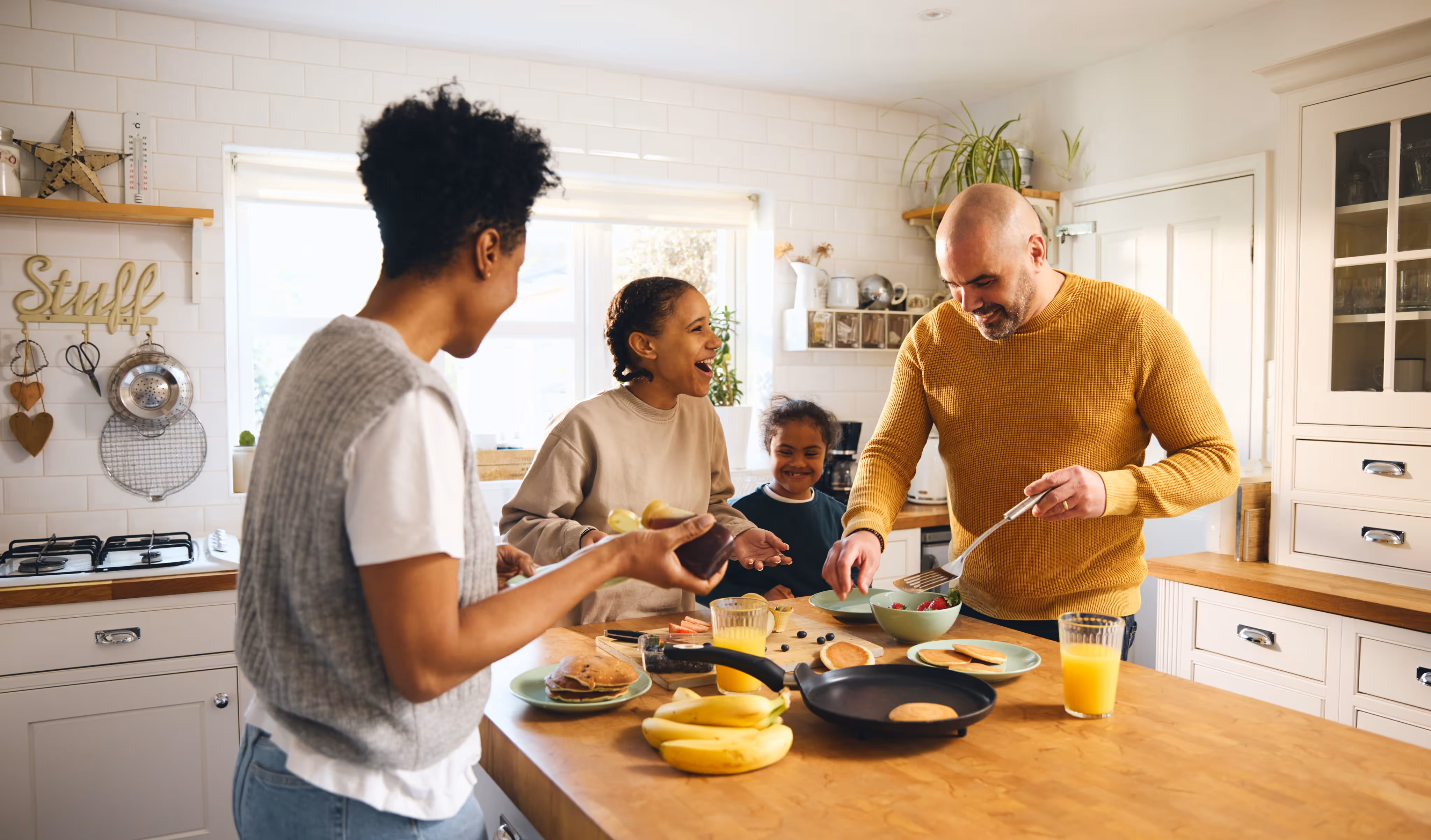 Family of four happily making pancakes together in a bright kitchen with fruit and orange juice.