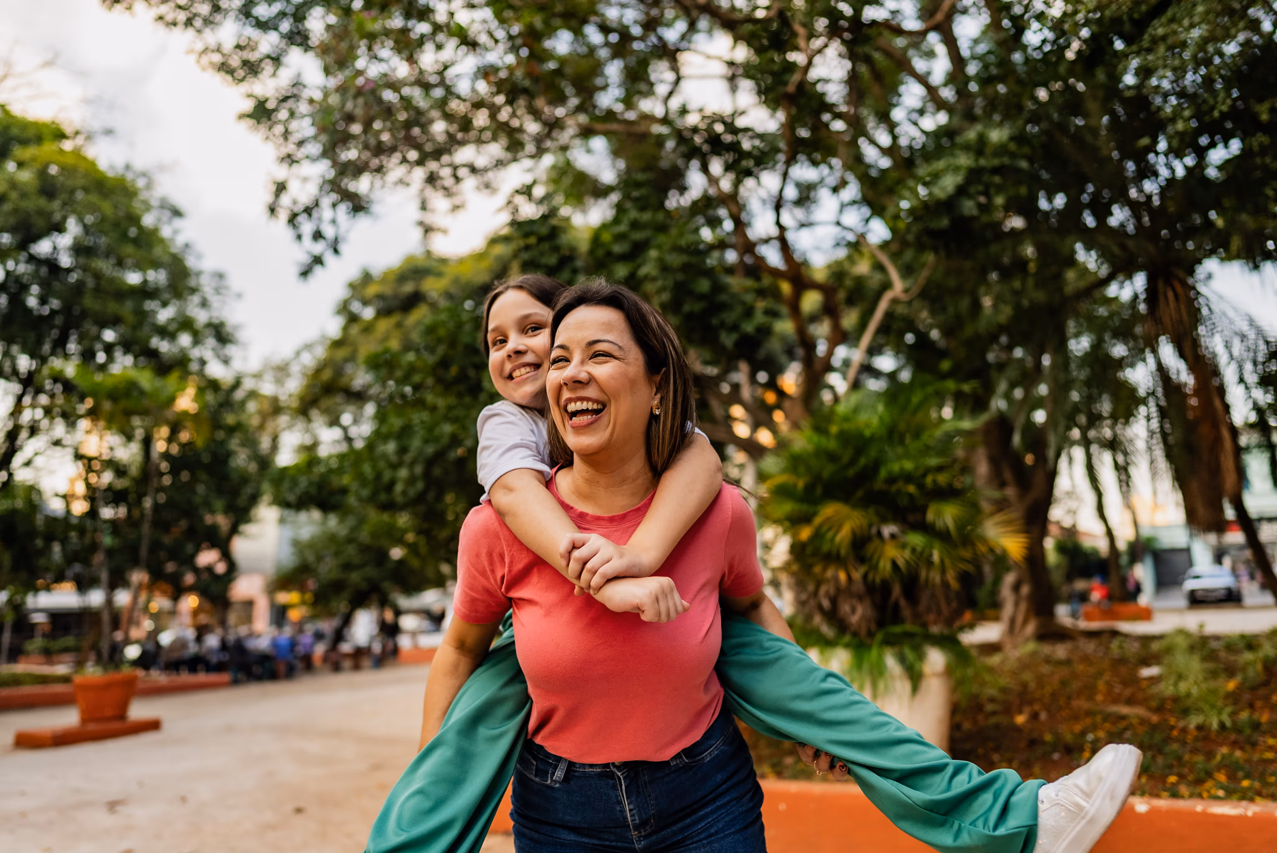 Smiling woman giving a piggyback ride to a laughing child in a park with green trees.