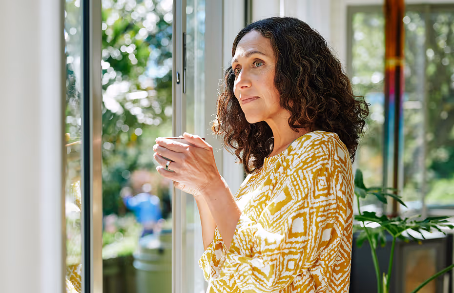 Woman in yellow patterned top holding a cup, looking thoughtfully out a window with plants inside.