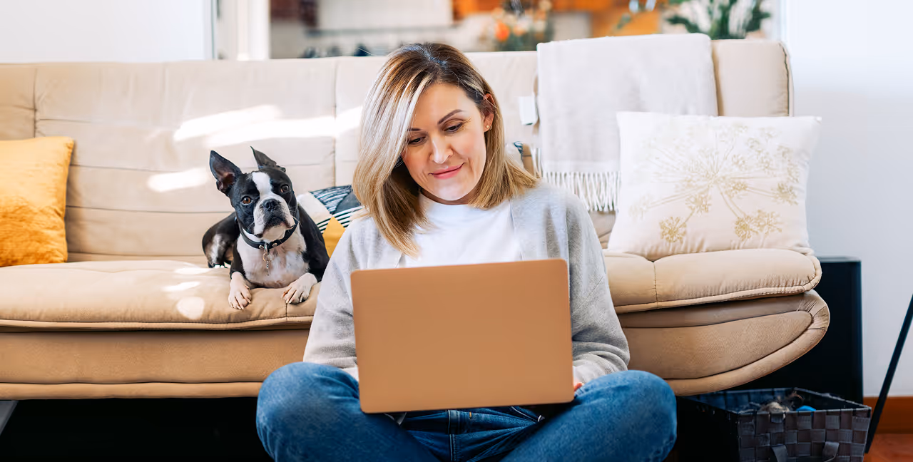 Woman sitting cross-legged on floor using laptop with a Boston terrier on the couch behind her.