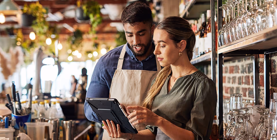 Two bar staff wearing aprons looking at a tablet together behind a bar counter with glasses.