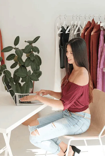 A young woman in a burgundy off-shoulder top and distressed jeans works on her laptop at a white table, surrounded by clothing racks and a potted plant, in a small business setting.