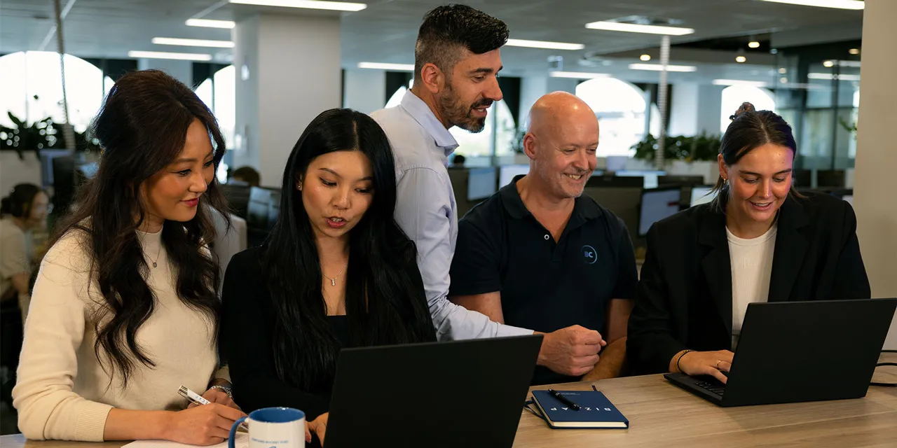 A team of five professionals, including two women and three men, collaborate over laptops in a modern office environment at small business loan company Bizcap's headquarters. 