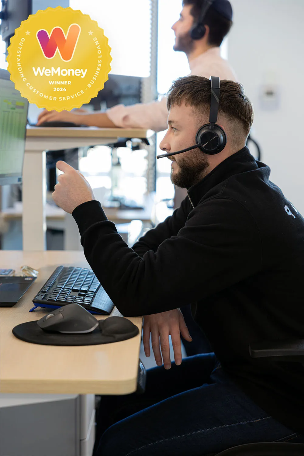 A member of the sales team at Bizcap wearing a black sweater and headset looking at a computer screen, discusses a business loan with customer.