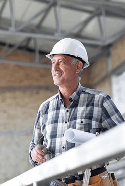 An older man in a checkered shirt and white hard hat smiles while holding architectural plans on a construction site.