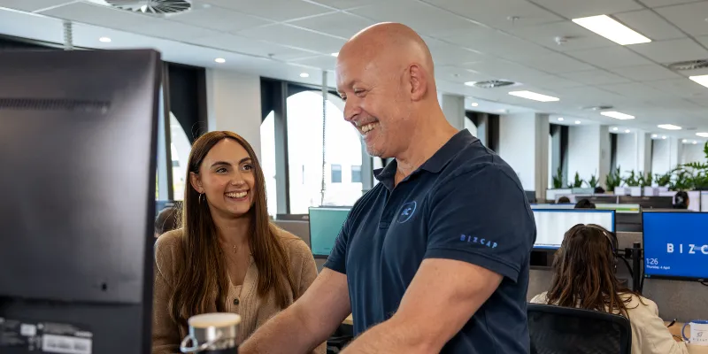 A broker in a blue shirt is assisting a smiling client at an office desk in a modern environment at the Bizcap office , both appearing engaged in their conversation.