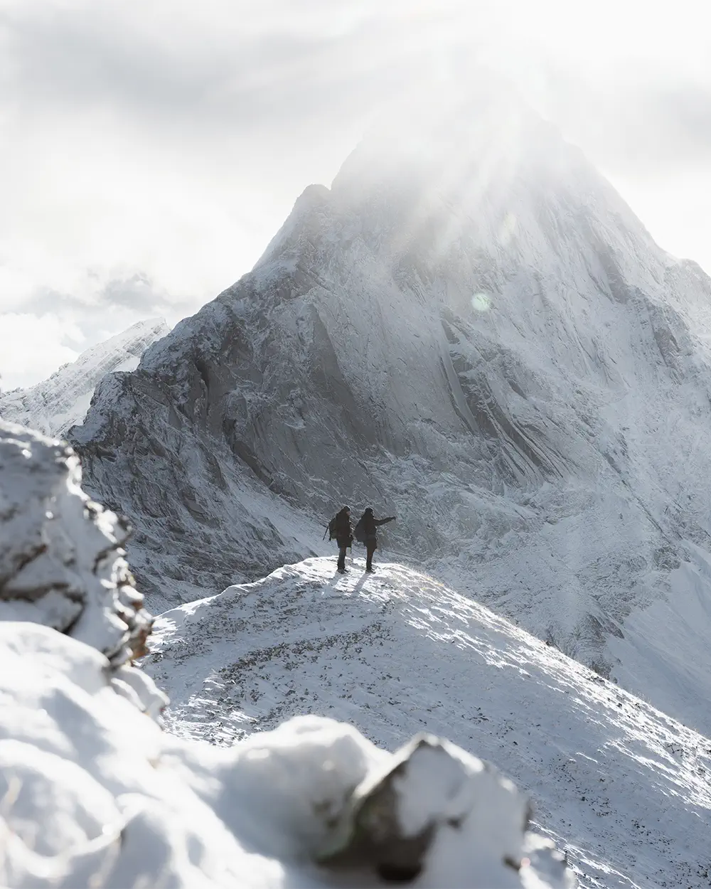 Two hikers stand on a snowy ridge with a towering snow-covered mountain in the background under a bright, cloudy sky.