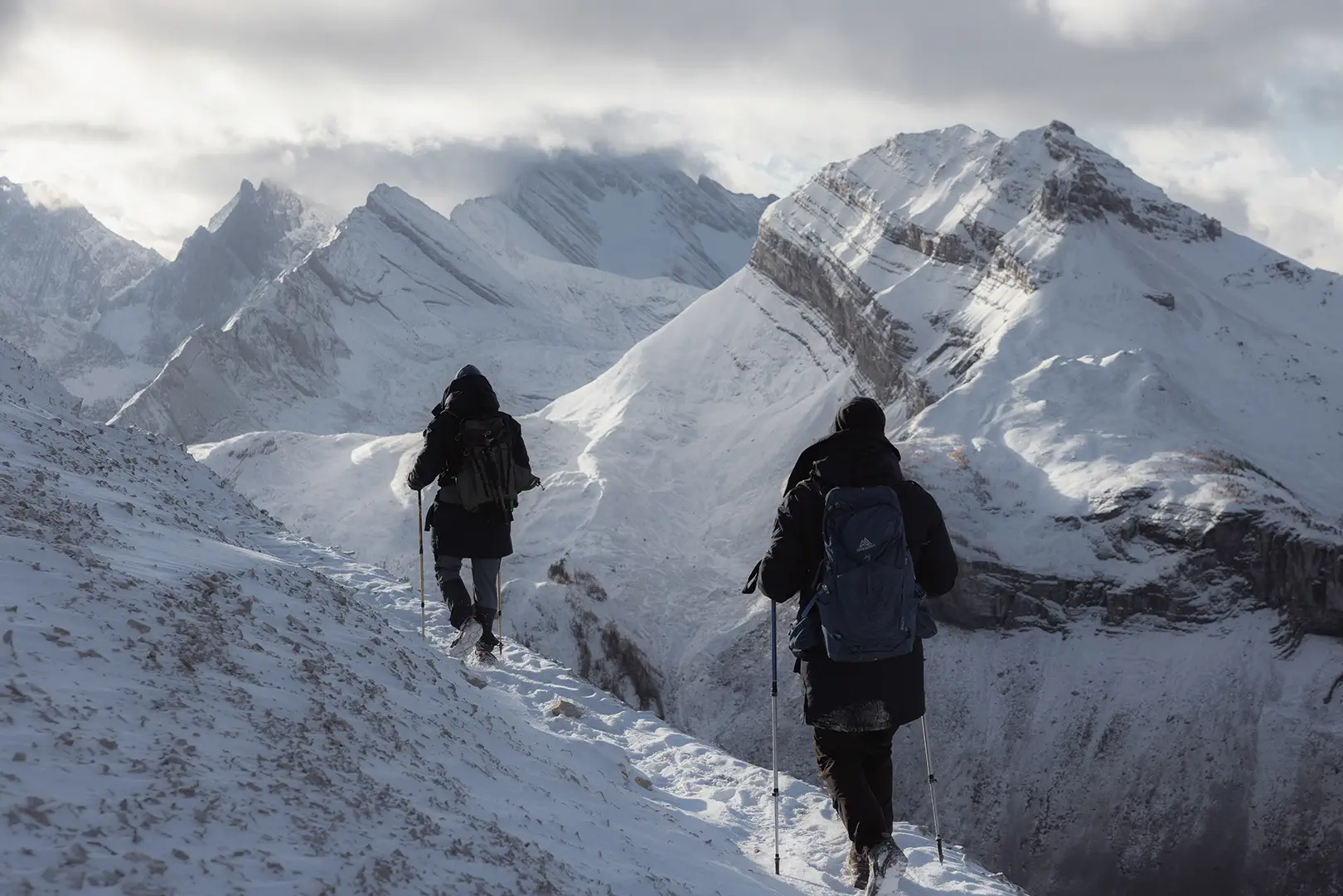 Two hikers wearing backpacks and winter gear walking on a snowy mountain trail with snow-covered peaks in the background.