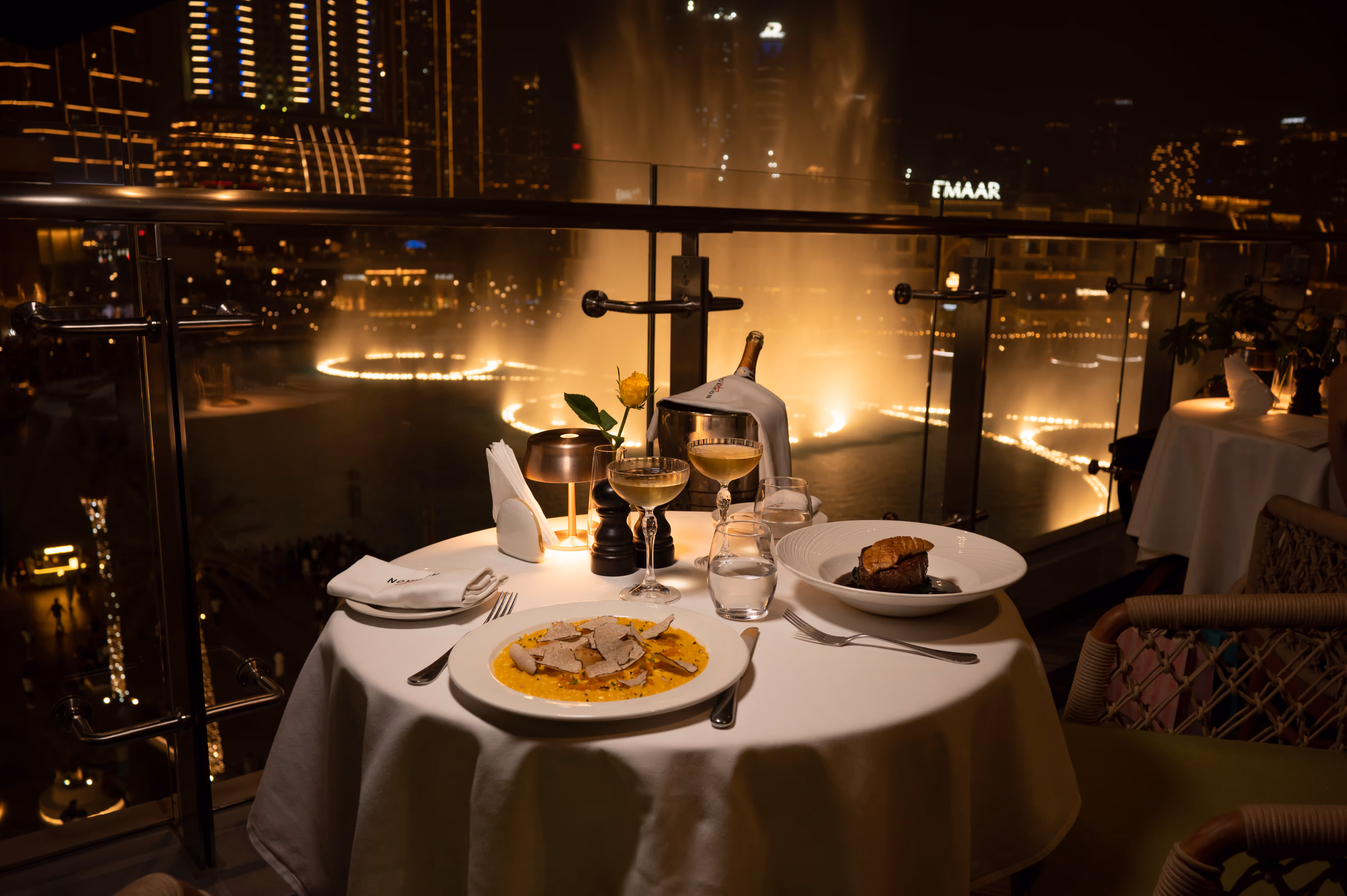 Elegant dinner setup on a balcony at night with two plated gourmet dishes, champagne glasses, and city lights with fountains in the background.