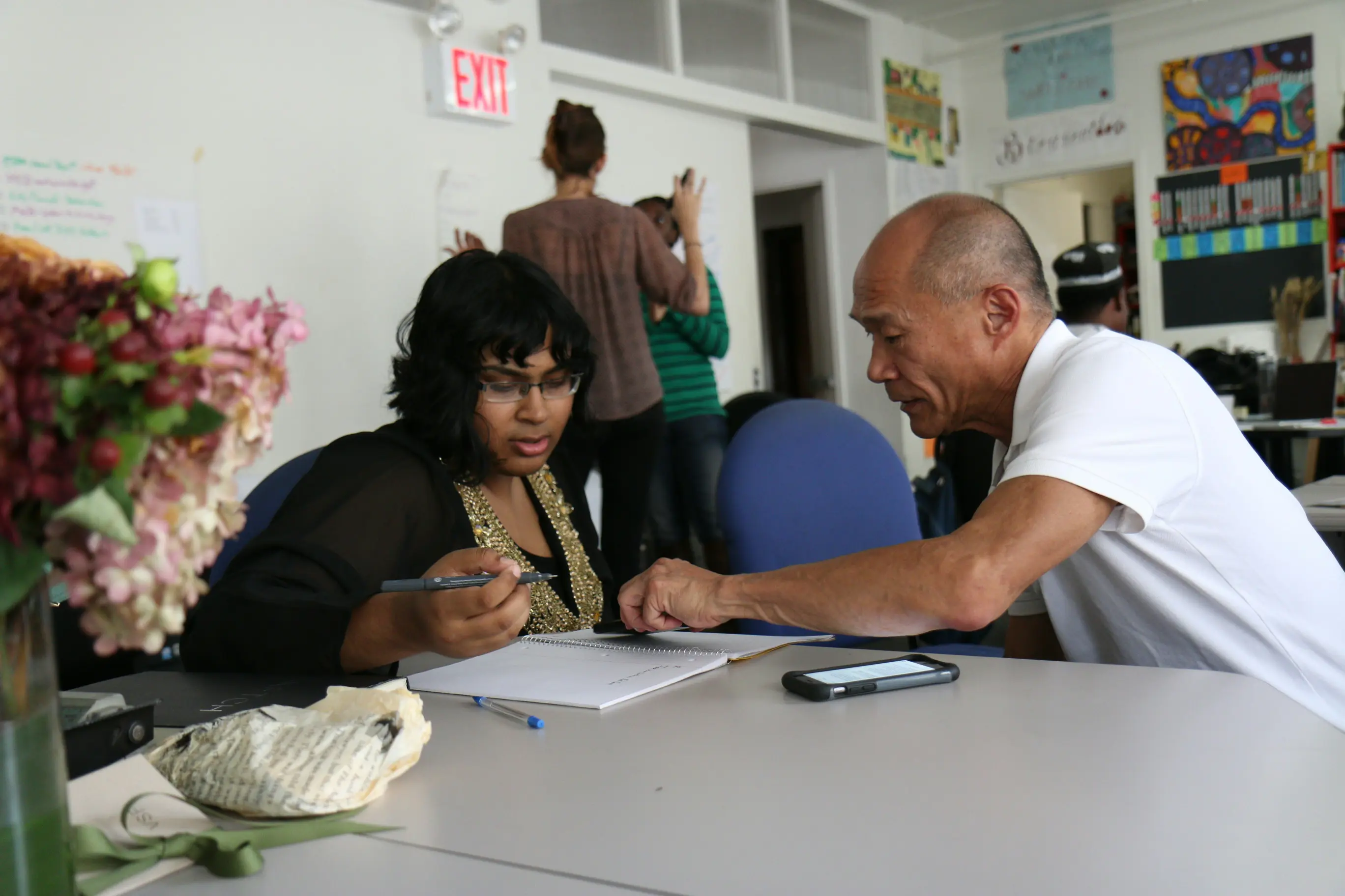 A student and teaching assistant sit together at a table. The teaching assistant, a man, shows the student, a woman, something in her notebook. 