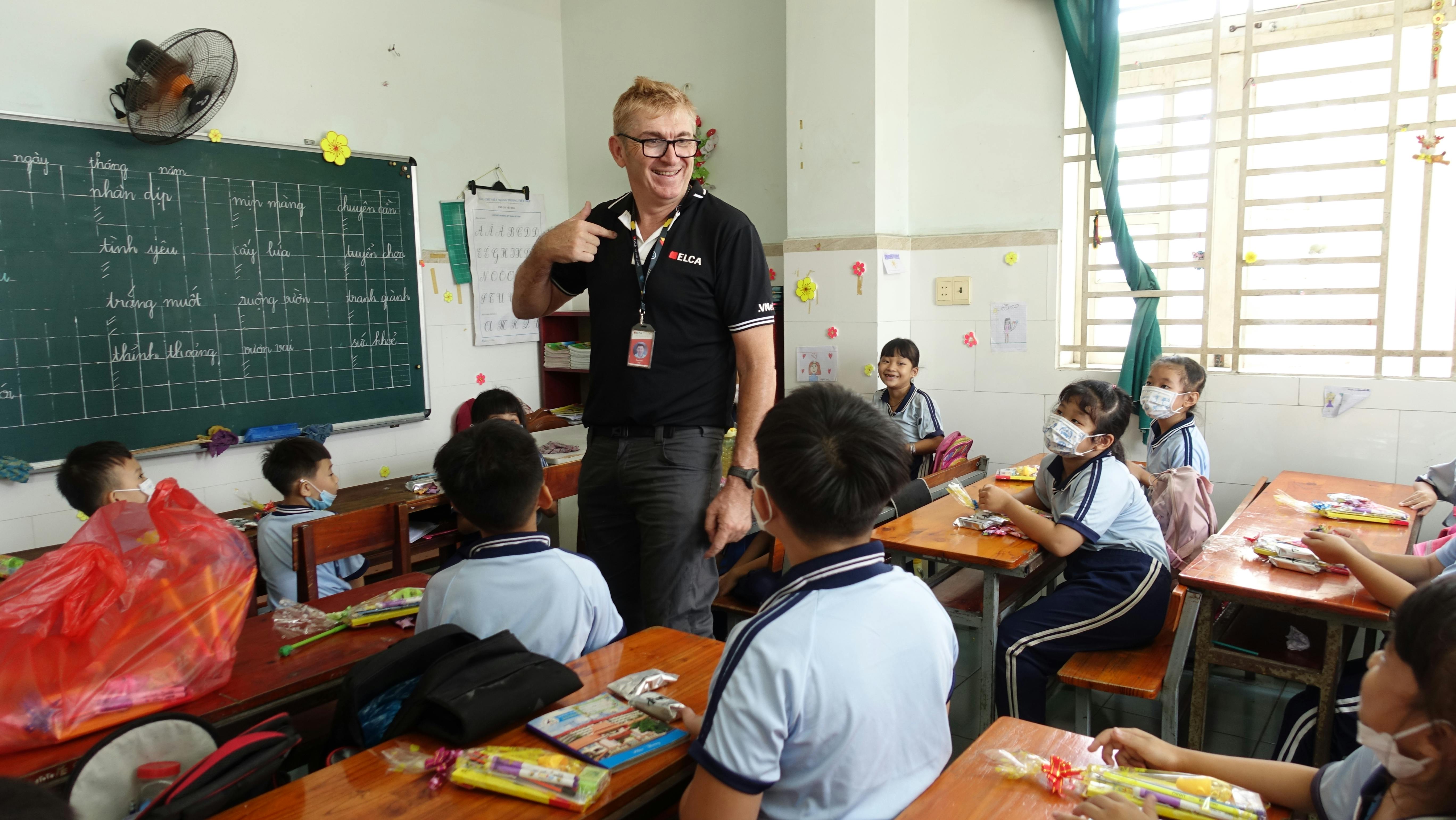 A cheerful male teacher stands at the front of a well-lit classroom, engaging primary students who sit at wooden desks with learning materials.