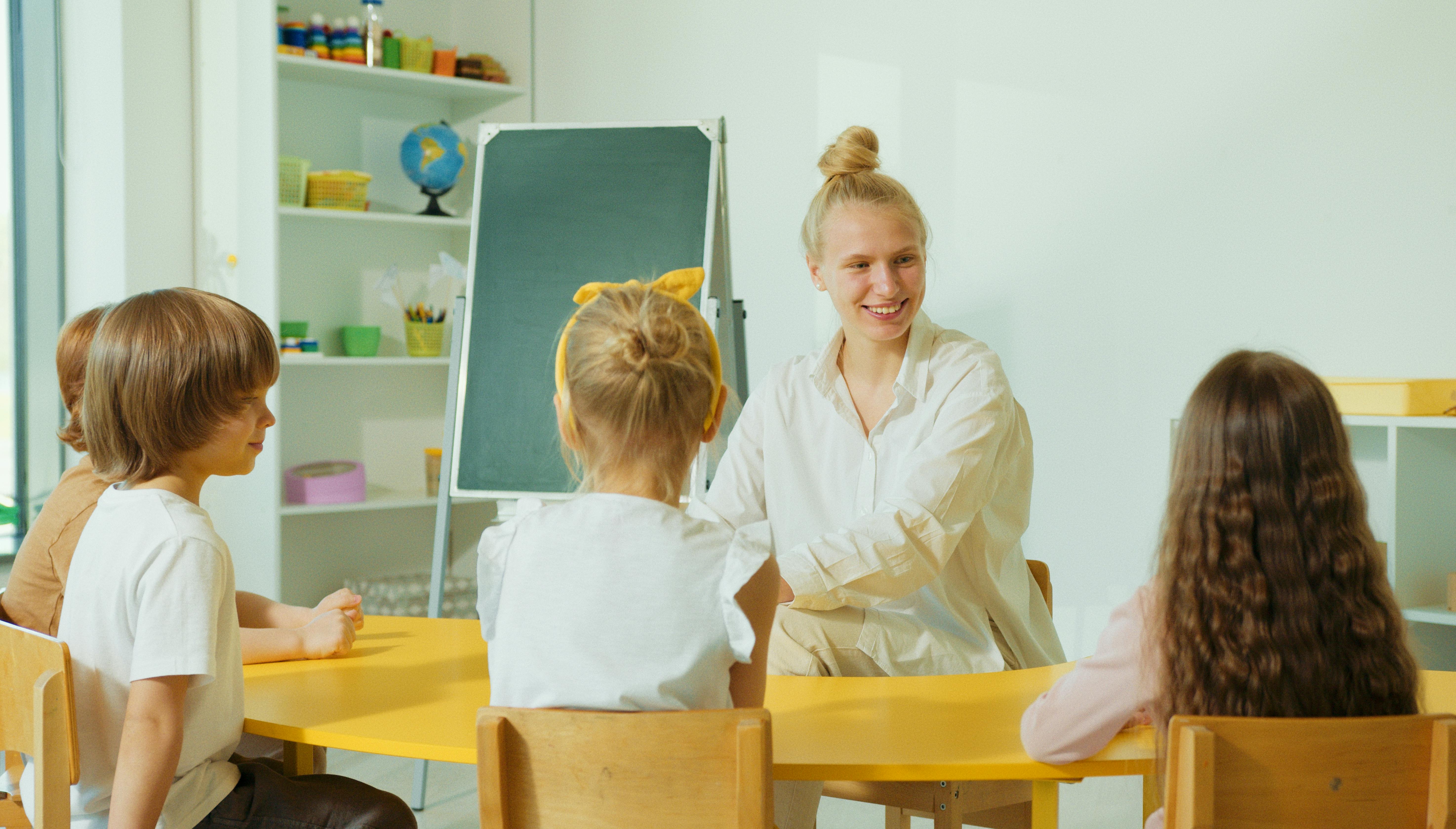 A smiling teacher sits at a table with three children in a bright classroom, with a small chalkboard and shelves of learning supplies behind them.