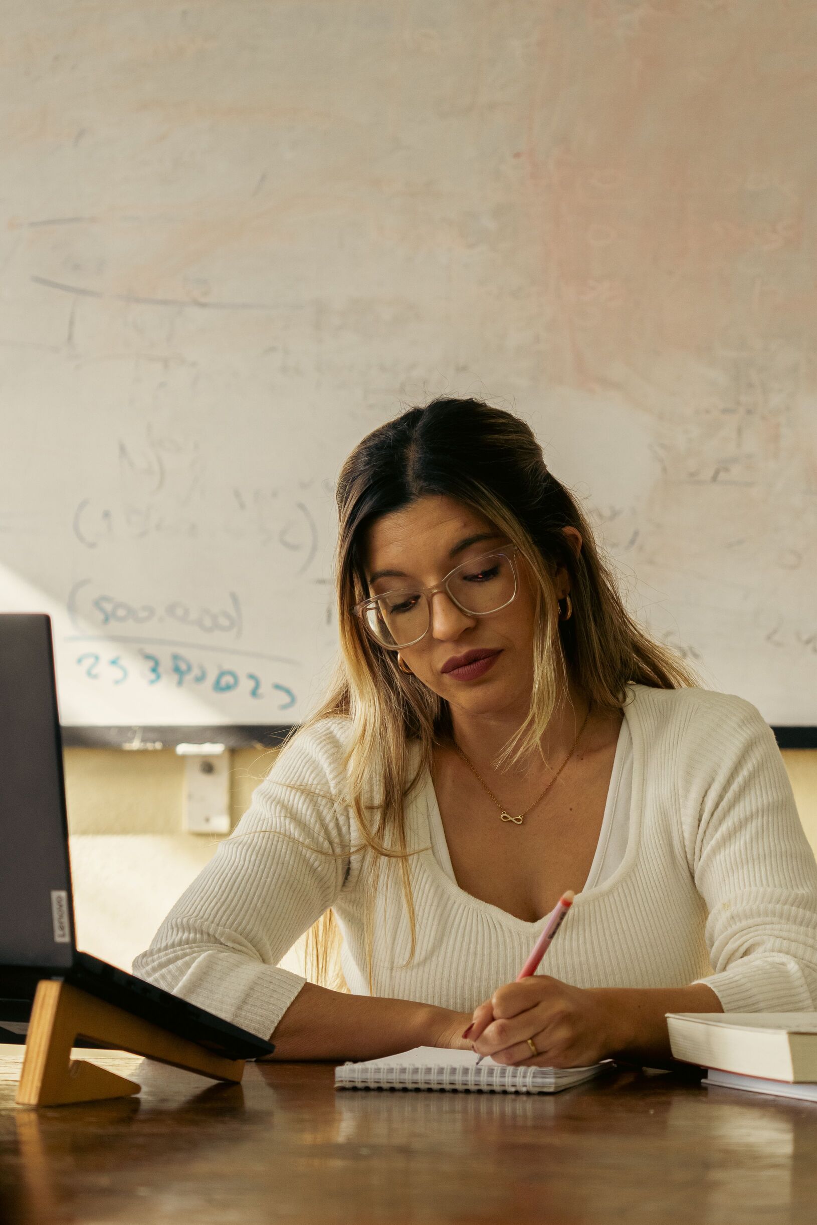 A woman wearing glasses writes thoughtfully in a notebook at a desk, with a laptop, books, and a whiteboard behind her.