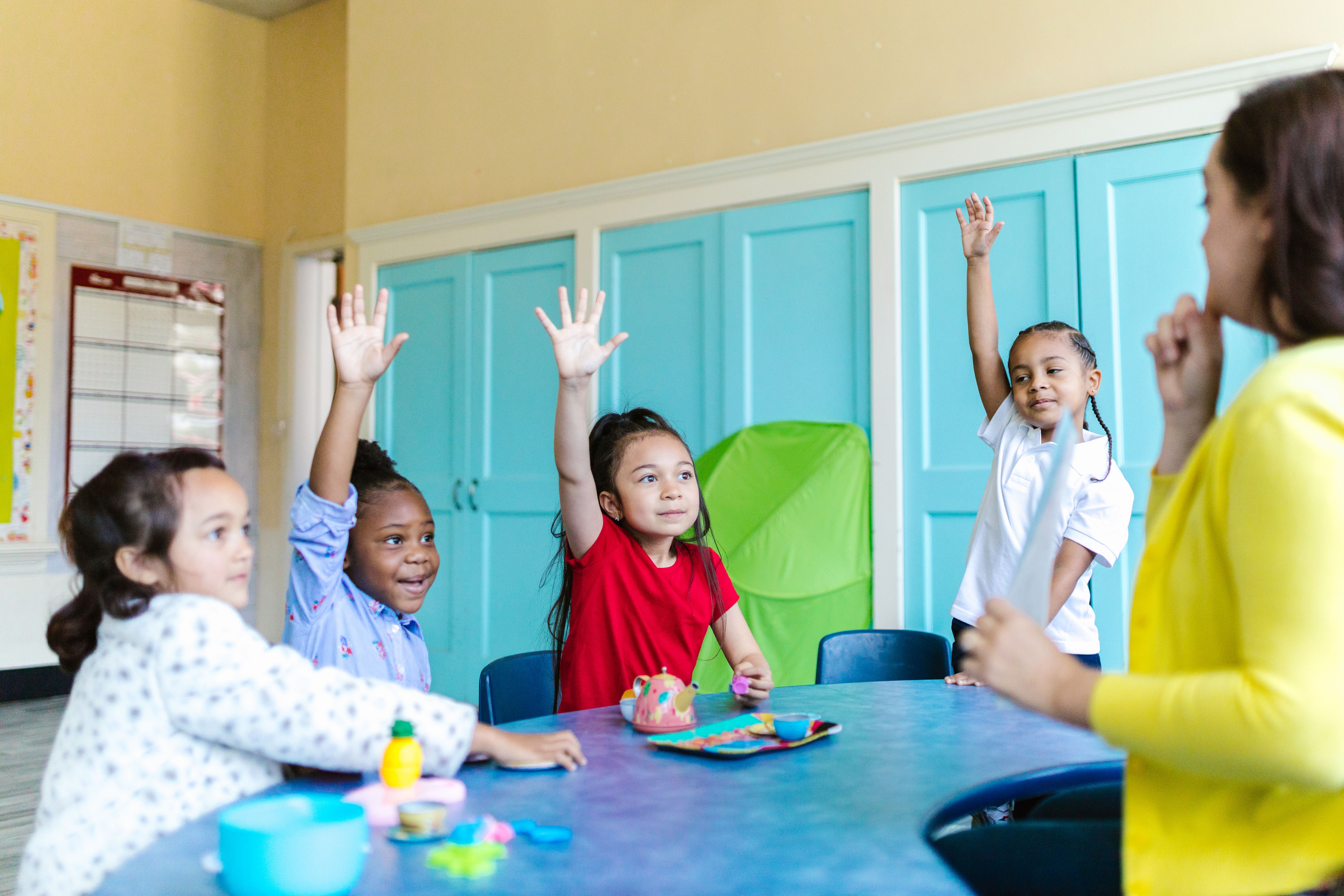 Children participating in a classroom activity while raising their hands and engaging with their teacher around a table in a colorful learning environment.