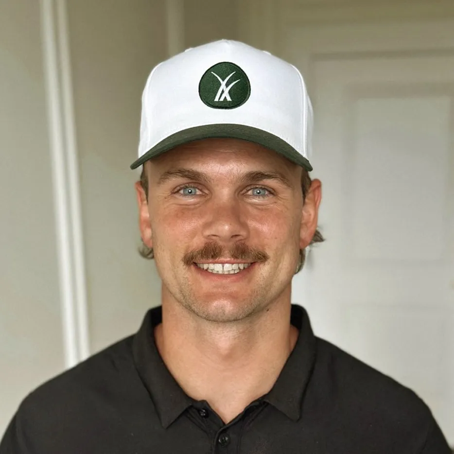 Smiling man with mustache wearing a white and green baseball cap and black shirt against a light background.