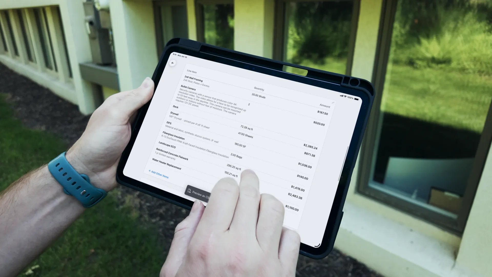 Close-up of a person holding a tablet outdoors displaying an itemized construction estimate with quantities and amounts for items like wall framing, bullet camera, drywall, EIFS, insulation, and concrete flatwork.