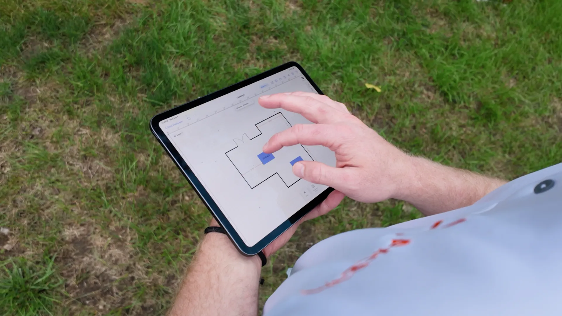A person using a tablet outdoors, holding it with one hand and using the other hand to interact with a floor plan displayed on the screen.