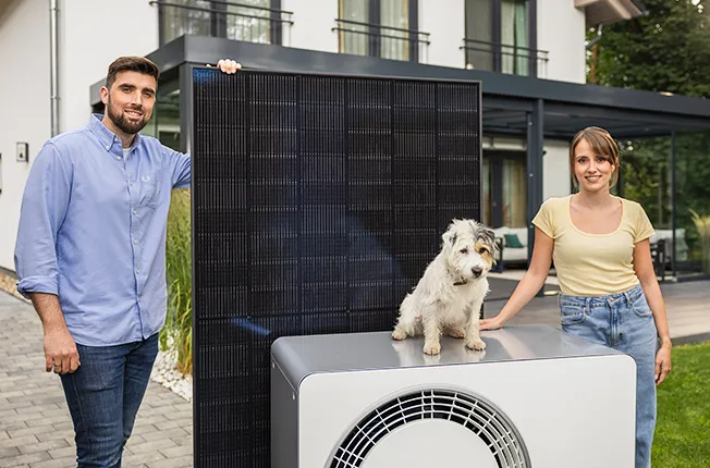 Enpal Kundenfamilie mit Solarmodul und Wärmepumpe vor dem Haus. Auf der Wärmepumpe sitzt ein kleiner Hund