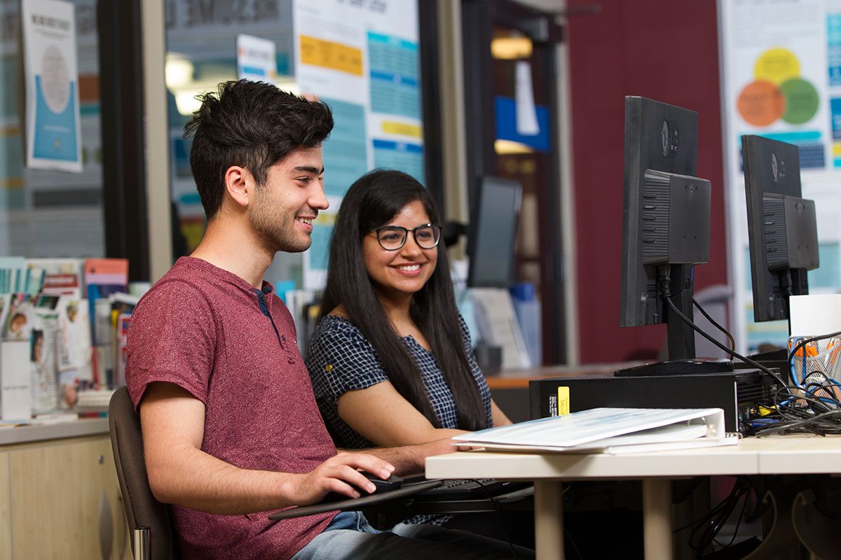 A young man and woman smiling while working together at a computer in a library or study area.