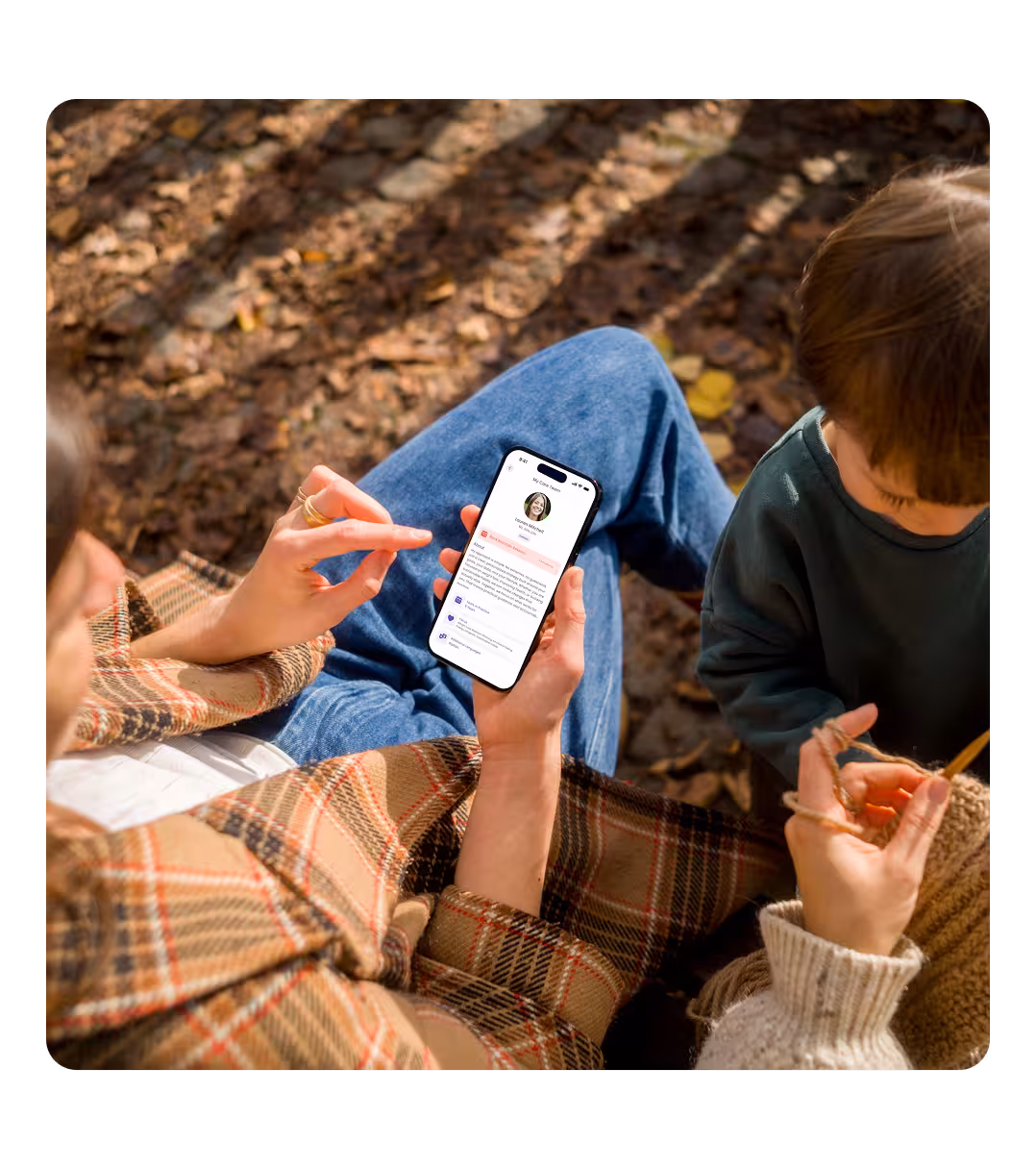 Person sitting outdoors on autumn leaves, holding a smartphone displaying a profile page, while another person beside them is knitting with yarn.