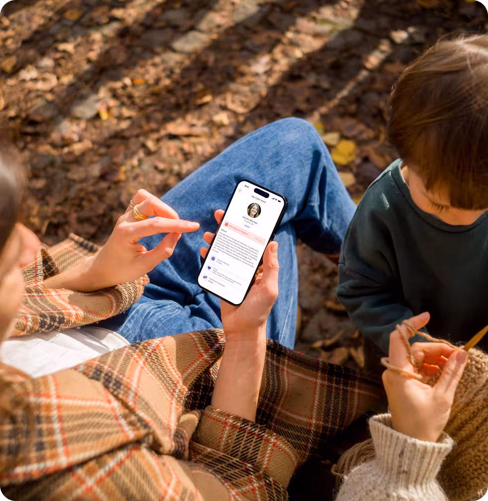 Person sitting outdoors on fallen leaves using a smartphone displaying a profile app, while another person knits nearby.