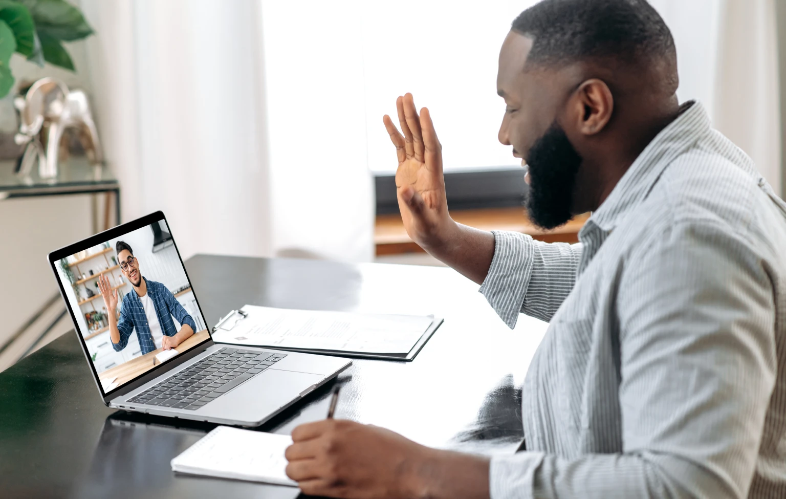 A man sits at a desk, smiling and waving during a virtual meeting with another person visible on his laptop screen. He holds a pen over a notebook, with documents and a clipboard nearby, suggesting an academic or support session in progress.