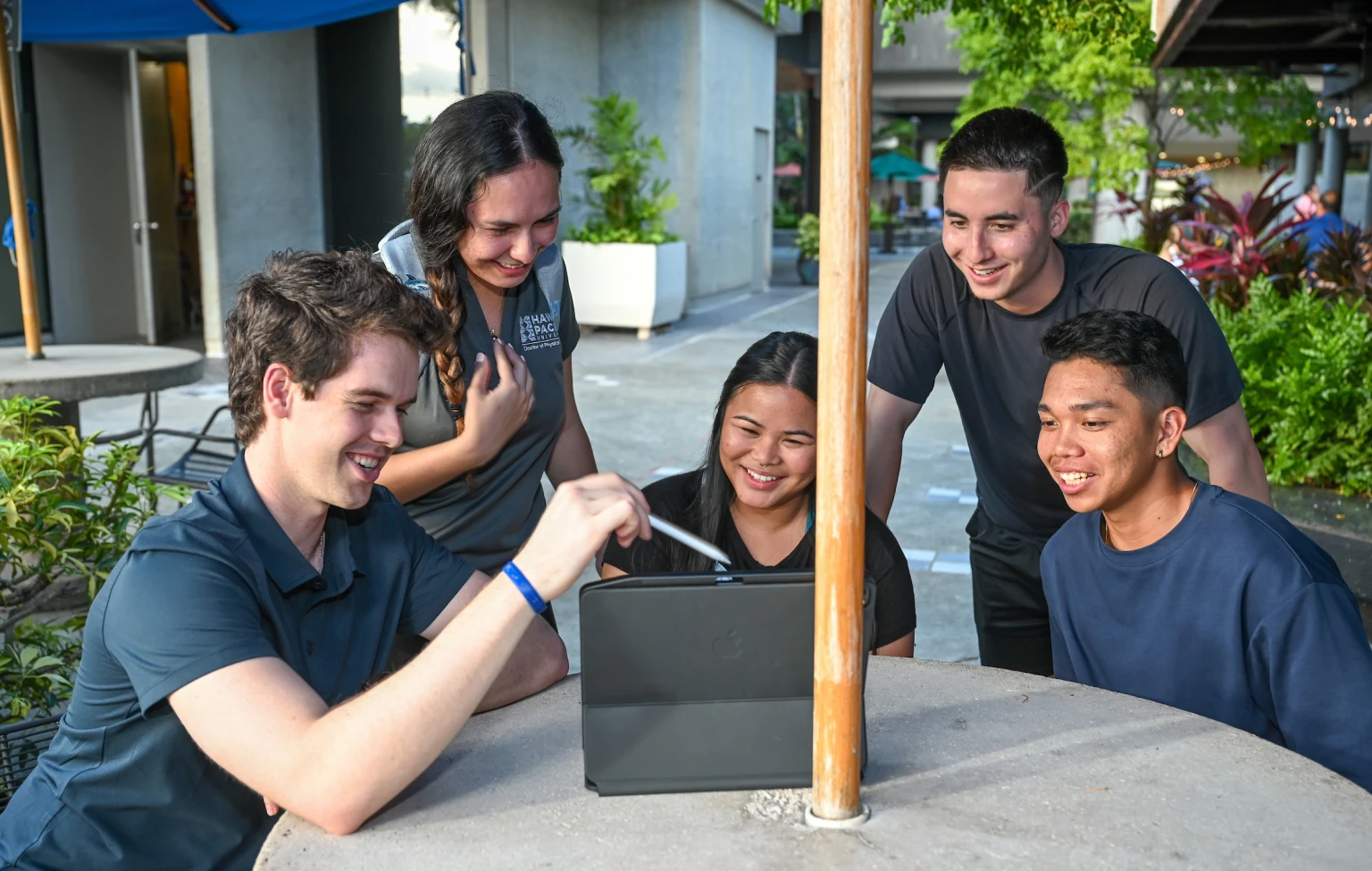 Five Hawai‘i Pacific University occupational therapy students gather around a tablet outdoors, smiling and collaborating on coursework.
