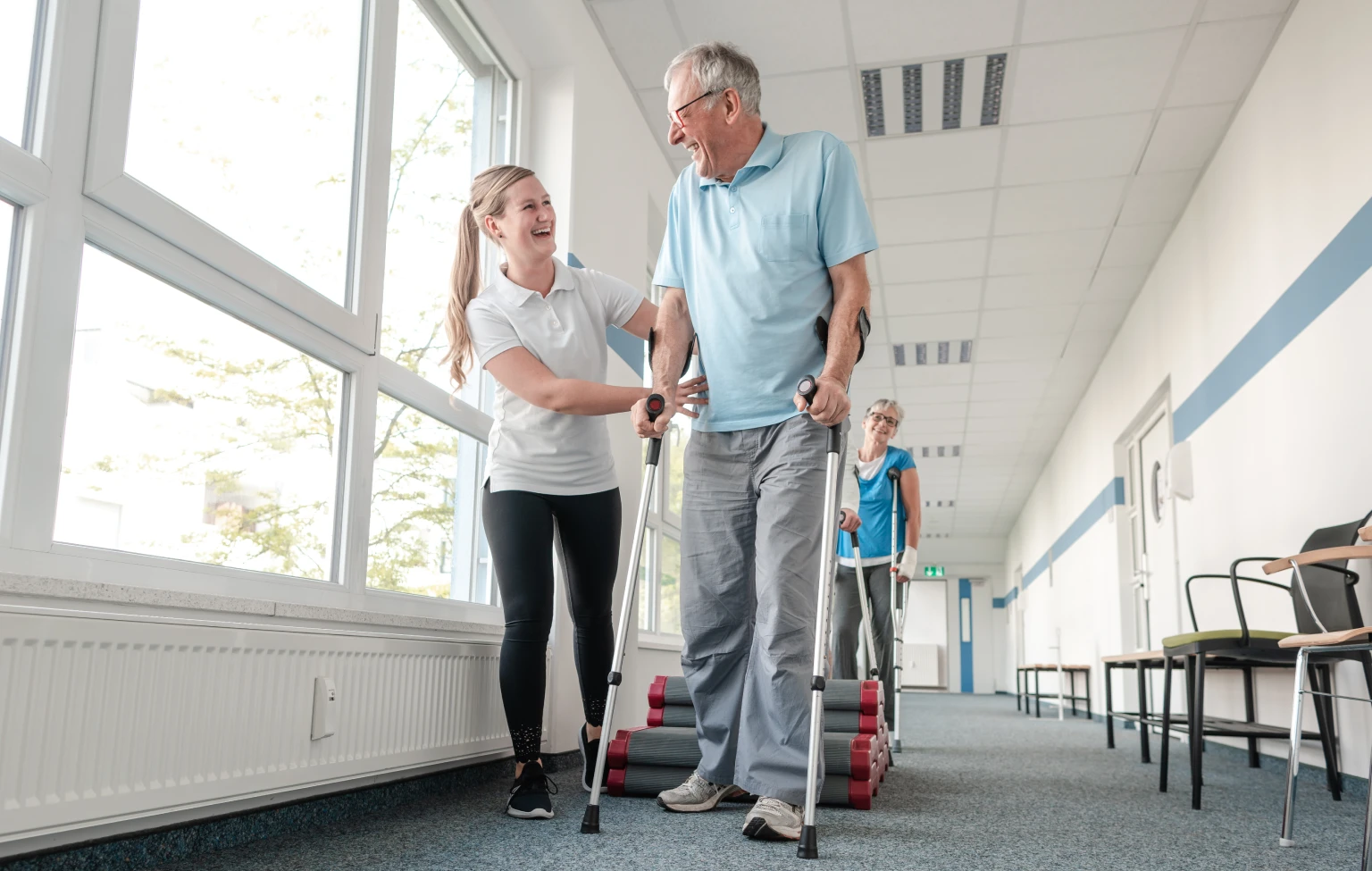 Physical therapy student supports older adult using crutches during rehabilitation session in a bright clinic hallway.