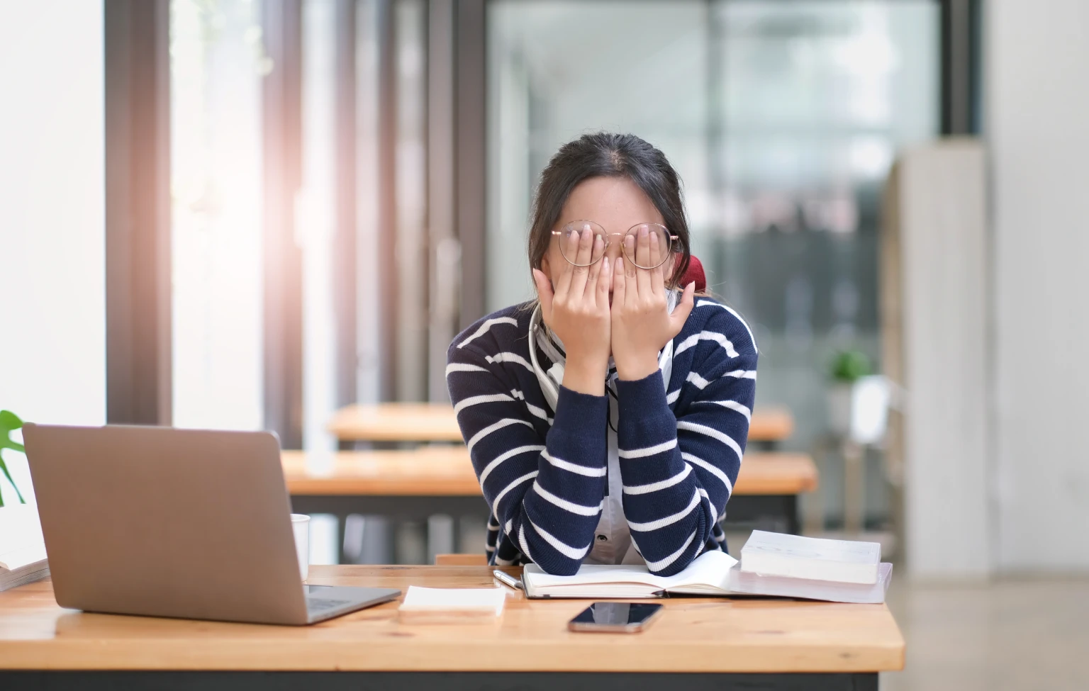 Stressed student in glasses covers face with hands while sitting at a desk with a laptop, books, and phone in a bright study space.