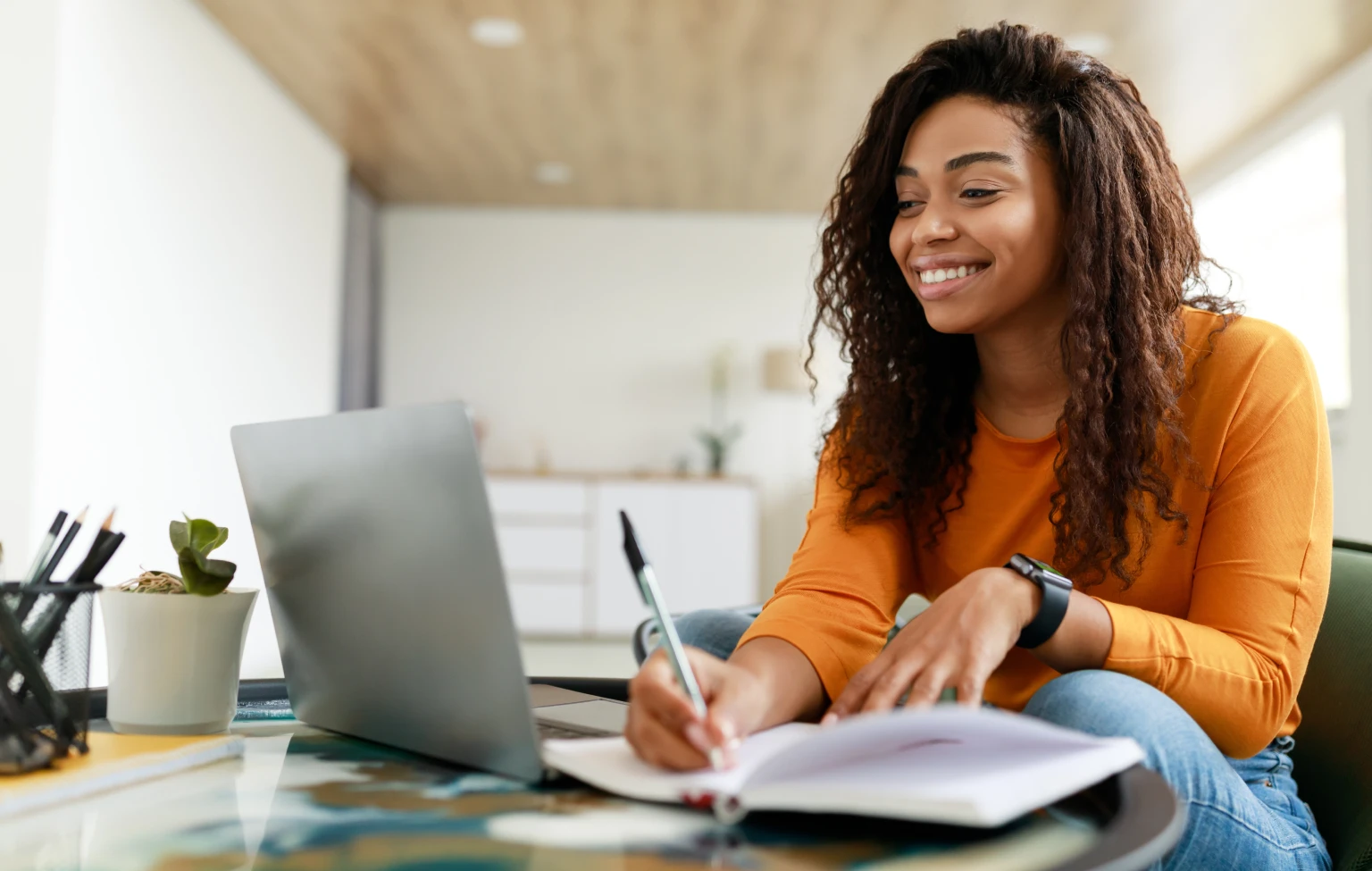 Student sitting at a desk smiling while taking notes and working on a laptop, illustrating online learning and flexible education.