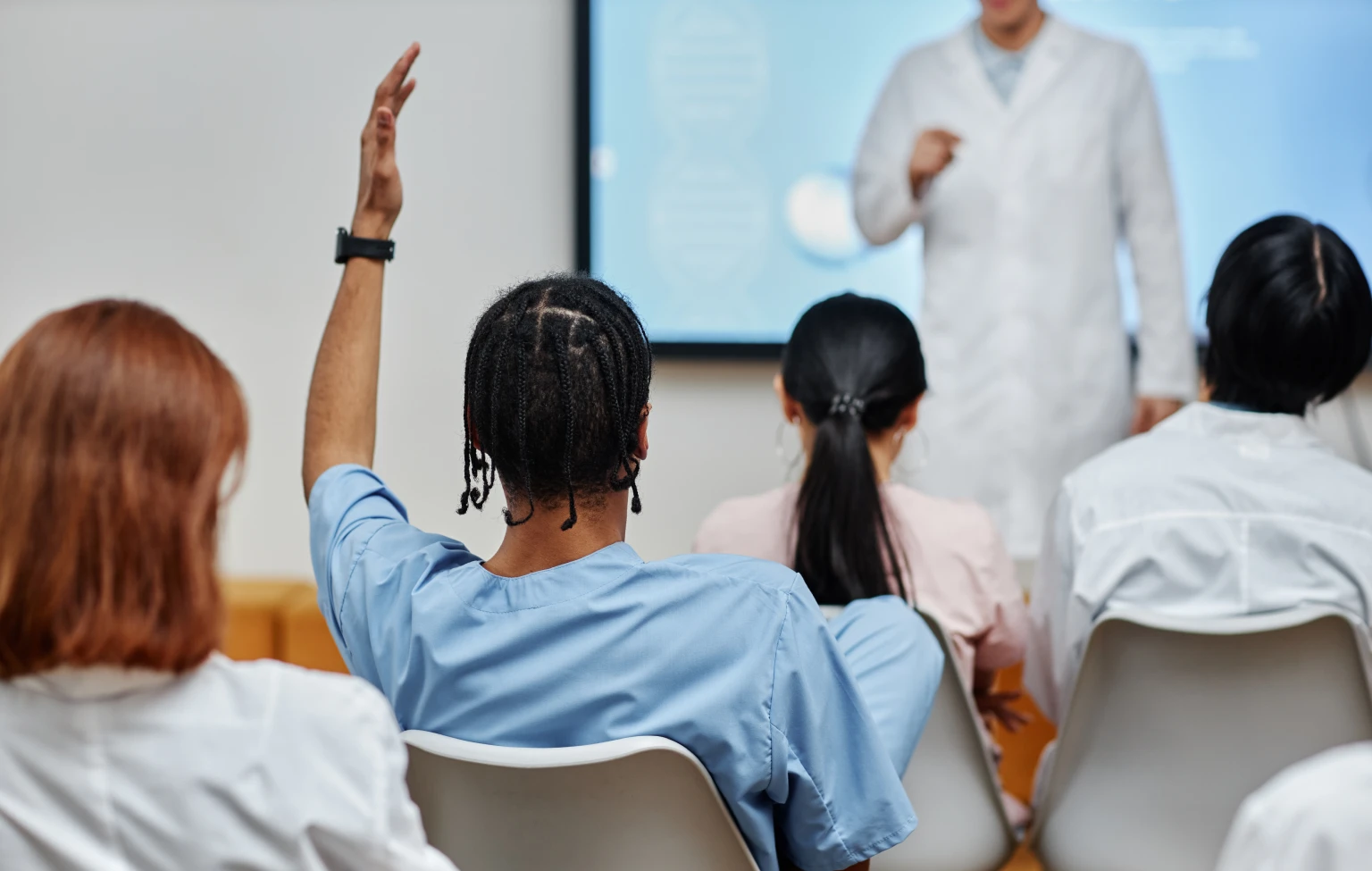 A group of health care students in a classroom listen to an instructor in a lab coat, with one student raising a hand to ask a question during a career-connected learning session.
