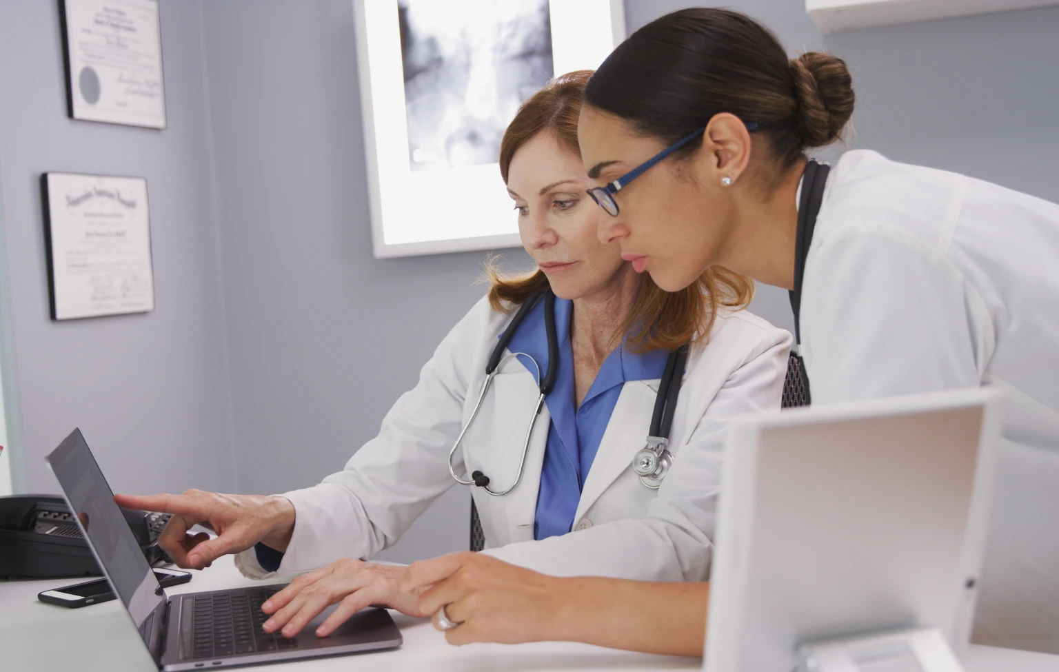 Two health care faculty members in lab coats review clinical information together on a laptop in a medical education setting.