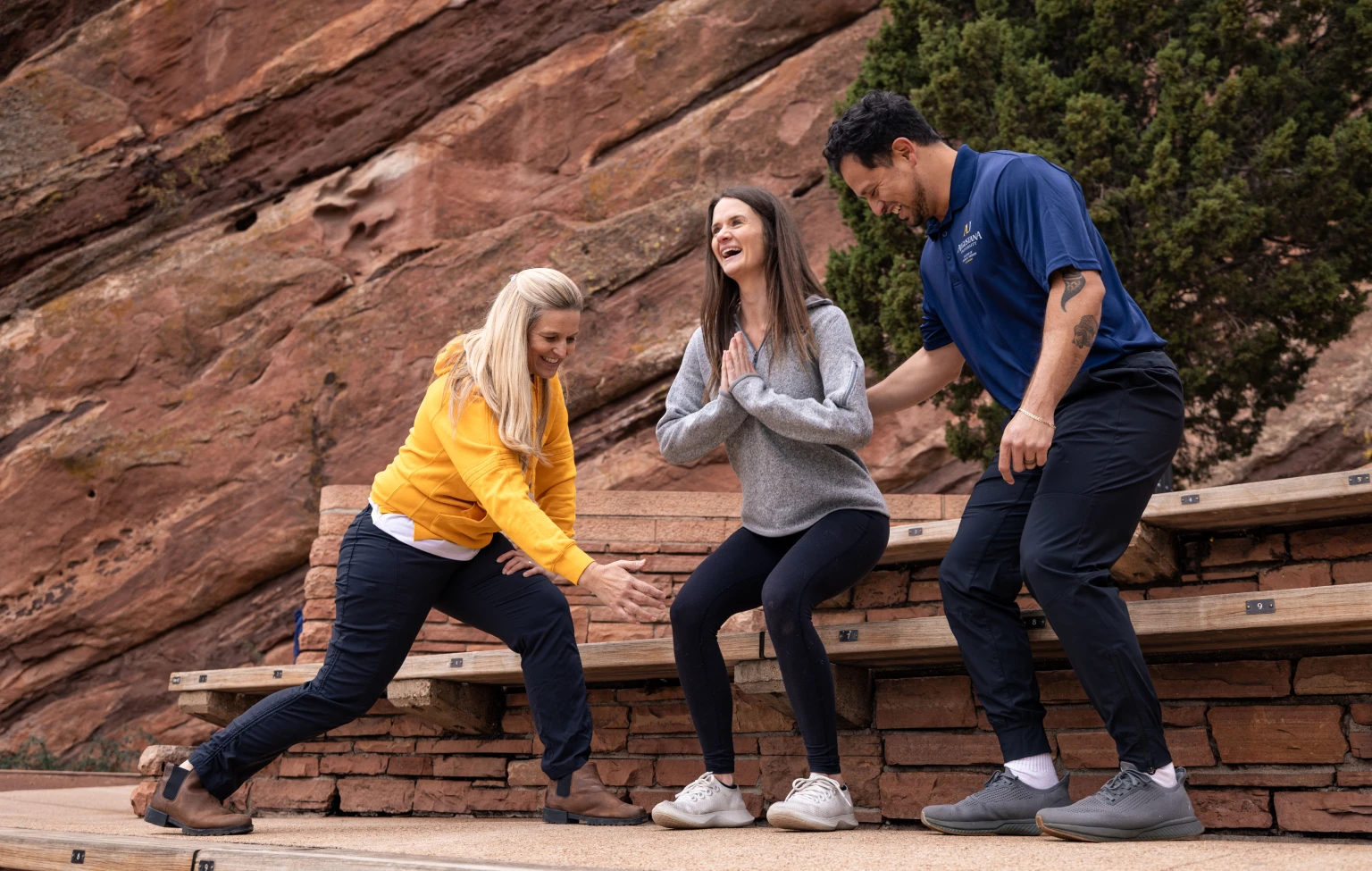 Three people participate in a hands-on physical therapy training exercise outdoors, practicing assisted movement and balance on bench seating near red rock formations.
