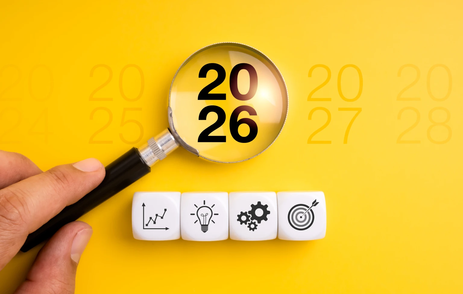 Hand holding a magnifying glass over the year ‘2026’ on a bright yellow background, with faint surrounding years and four white blocks showing icons for growth, innovation, systems, and target/strategy.
