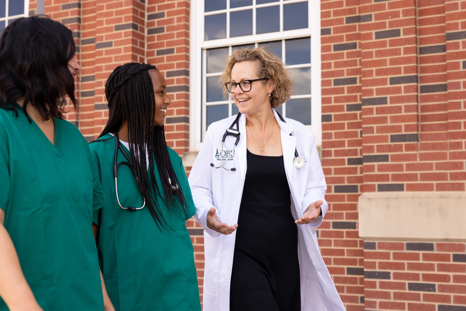 Physician associate instructor in a white coat speaks with two students in green scrubs outside a campus building.