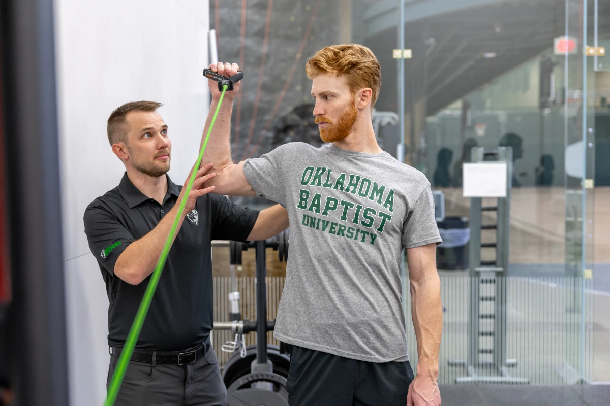 Physical therapy student performs resistance band shoulder exercise with instructor guidance at Oklahoma Baptist University.