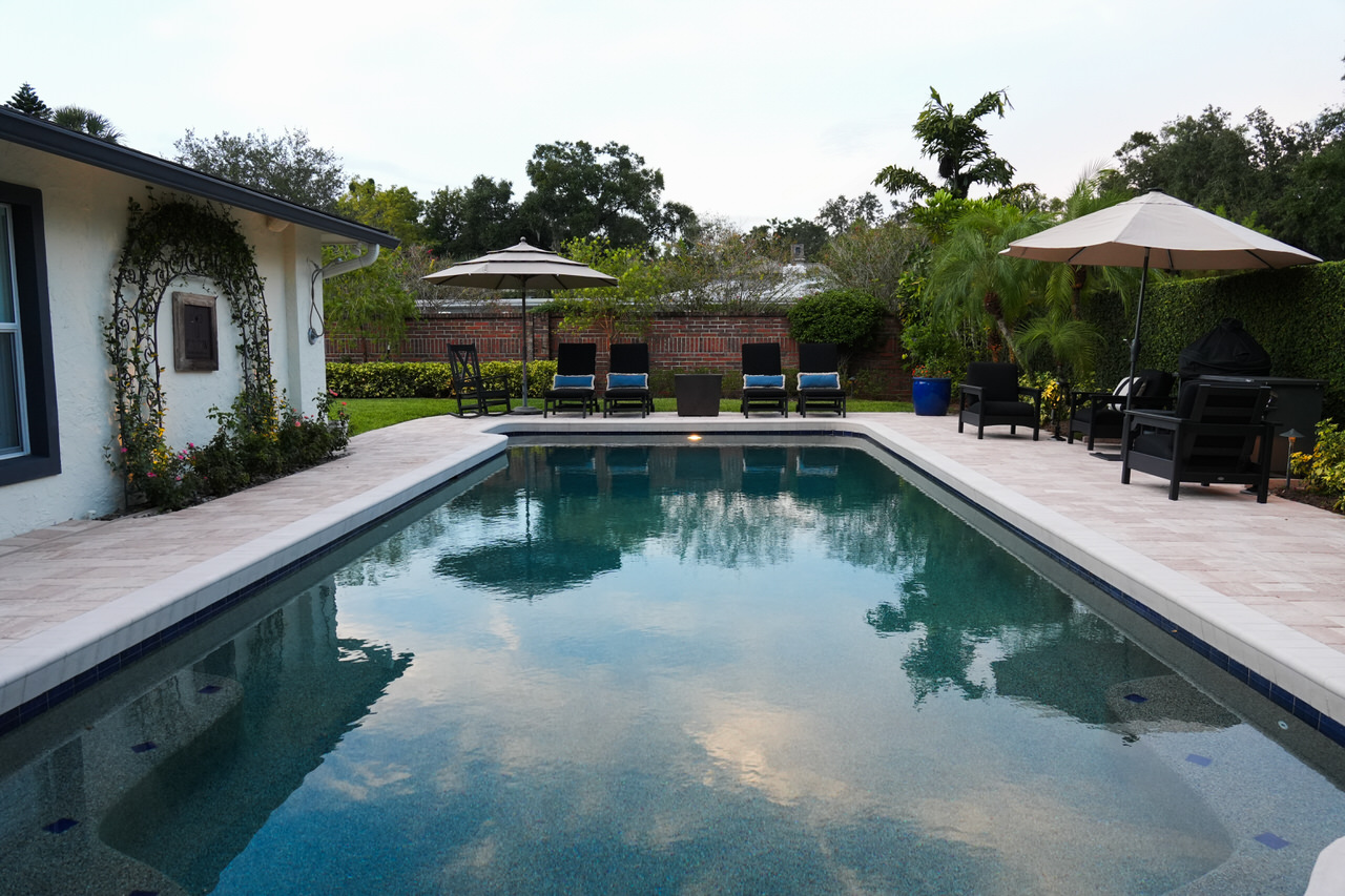 Rectangular outdoor swimming pool with patio chairs, umbrellas, and a brick wall in a backyard garden.