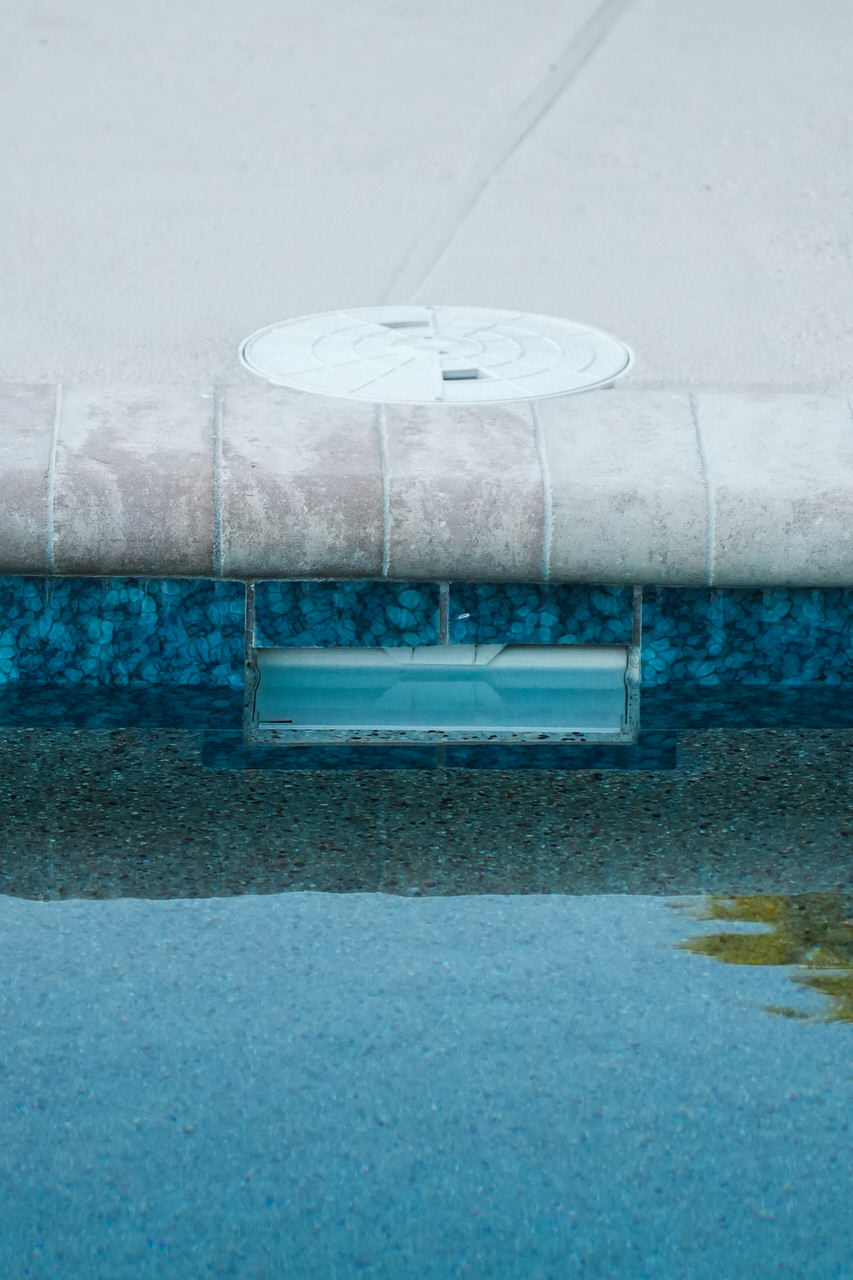 Close-up of a swimming pool edge showing blue-tiled waterline and beige pool coping with a round drain cover on the deck.