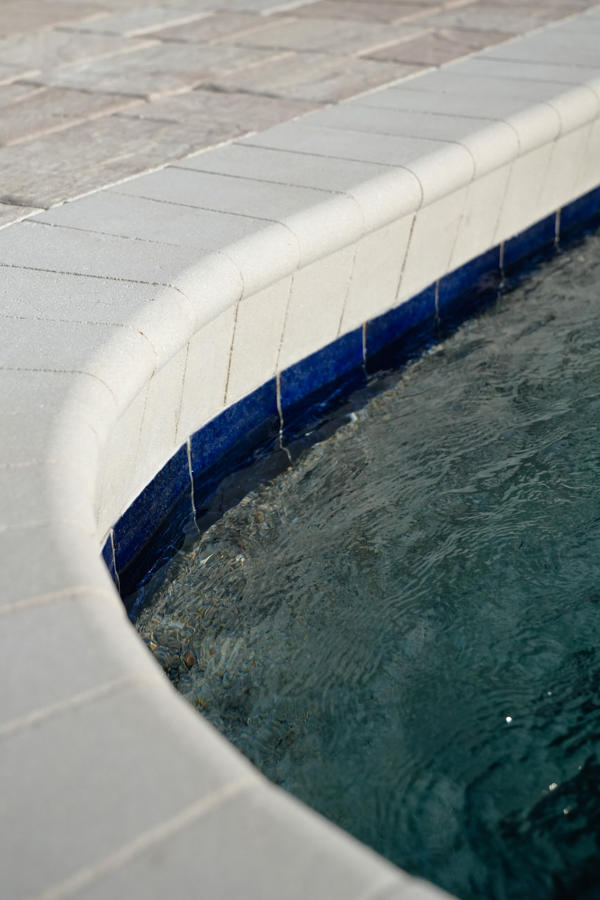 Curved edge of a swimming pool with clear water and blue tile trim beneath white stone coping and surrounding stone pavement.
