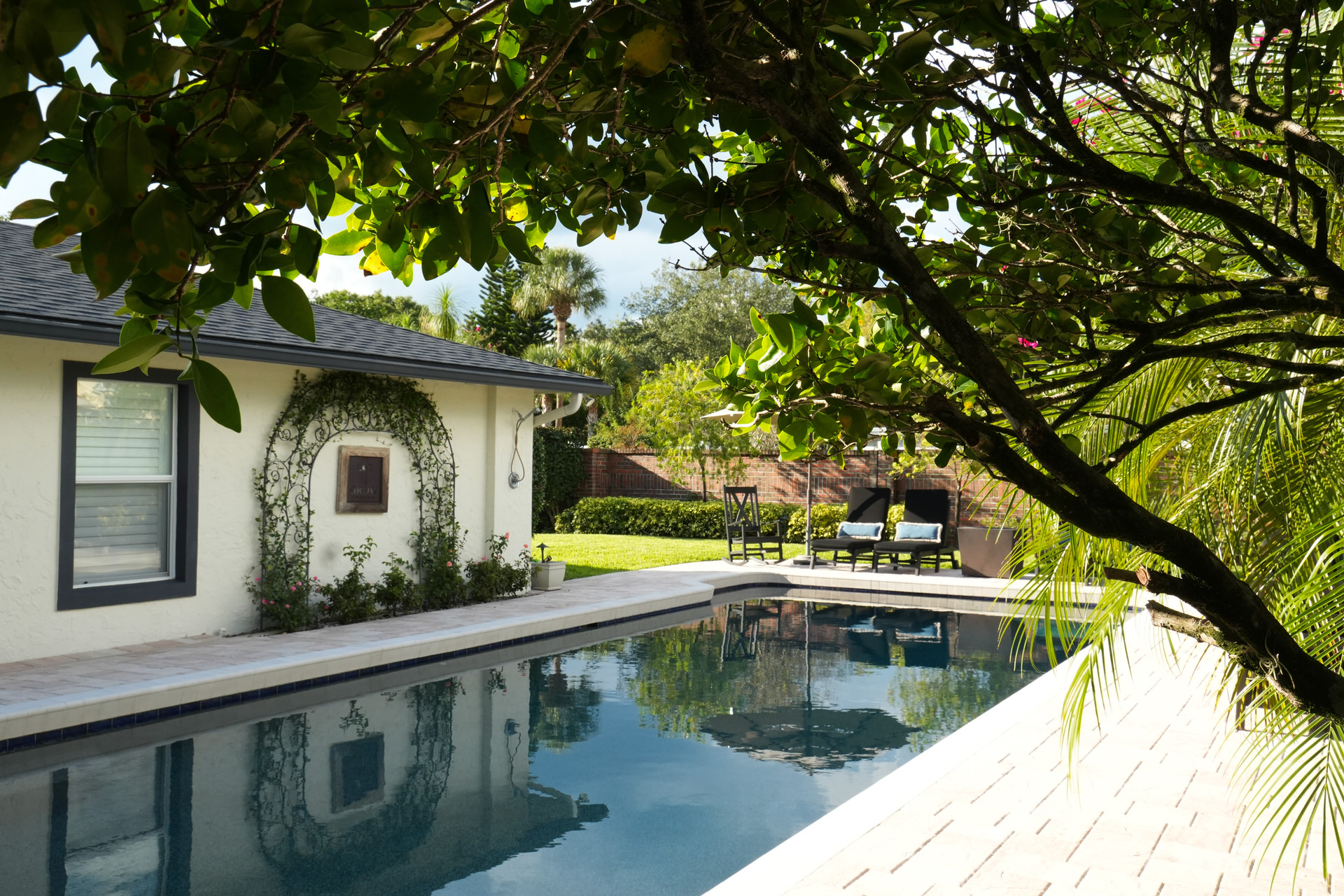 Backyard inground pool with patio, lounge chairs, and green trees surrounding a beige house.