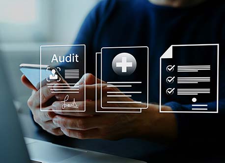 Man sitting at desk holding mobile phone with document icons hovering infront of him with one icon displaying the text audit