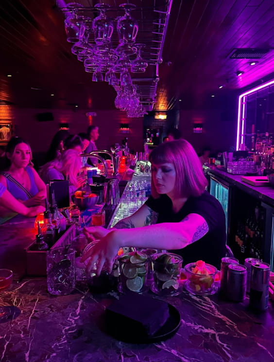 A woman stands behind a bar illuminated by purple lighting, creating a vibrant and inviting atmosphere. 