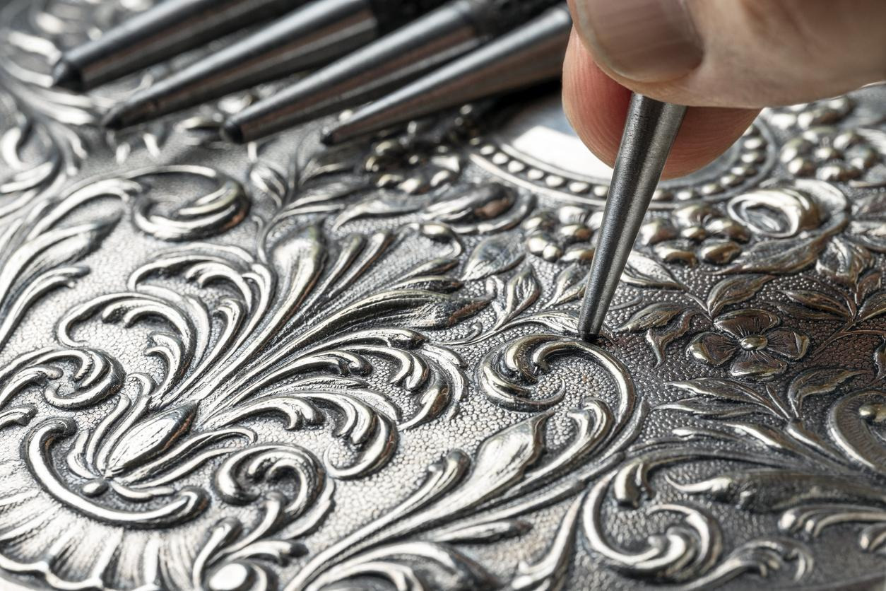 A person uses a silver tool to engrave a design onto a silver plate, showcasing intricate craftsmanship.  