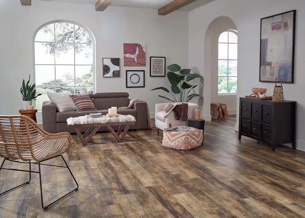 A contemporary living room featuring luxury vinyl plank flooring with rich brown tones and realistic wood textures. The space is styled with a mix of modern and bohemian decor, including a large arched window, rattan furniture, and an array of indoor plants, creating a warm and inviting atmosphere.